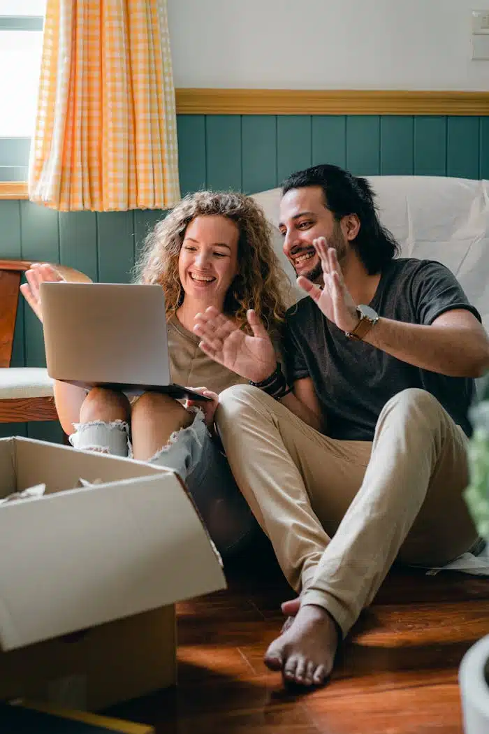 Full body happy smiling diverse couple in casual wear sitting on floor near cardboard boxes in new apartment and chatting via netbook